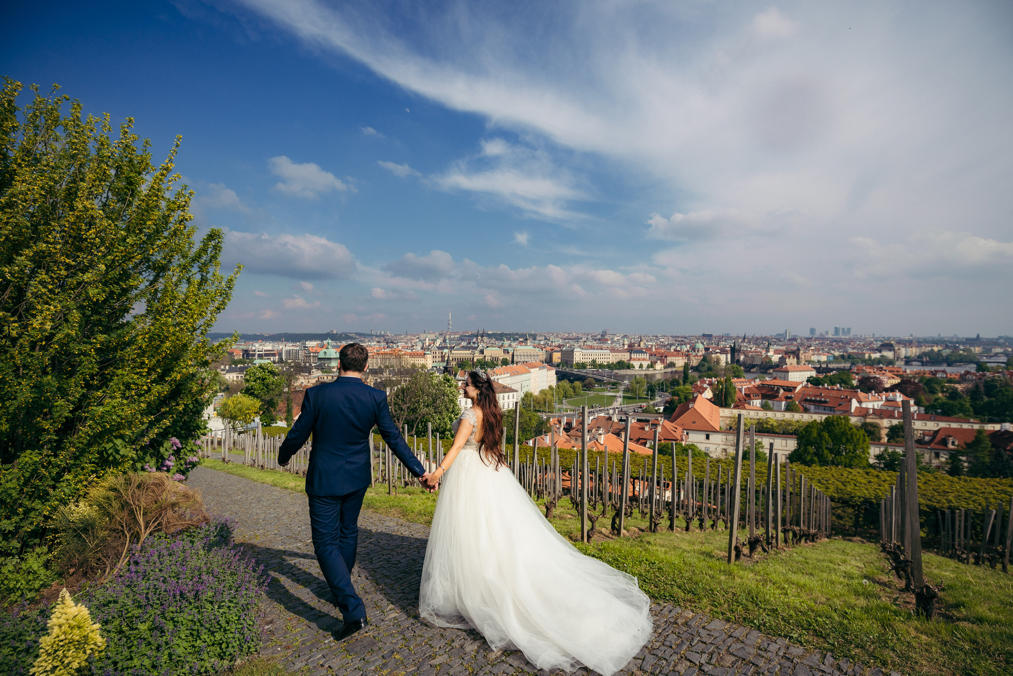 The back view of the newlyweds holding hands and walking along the paving road under the beautiful sky. Prague panorama
