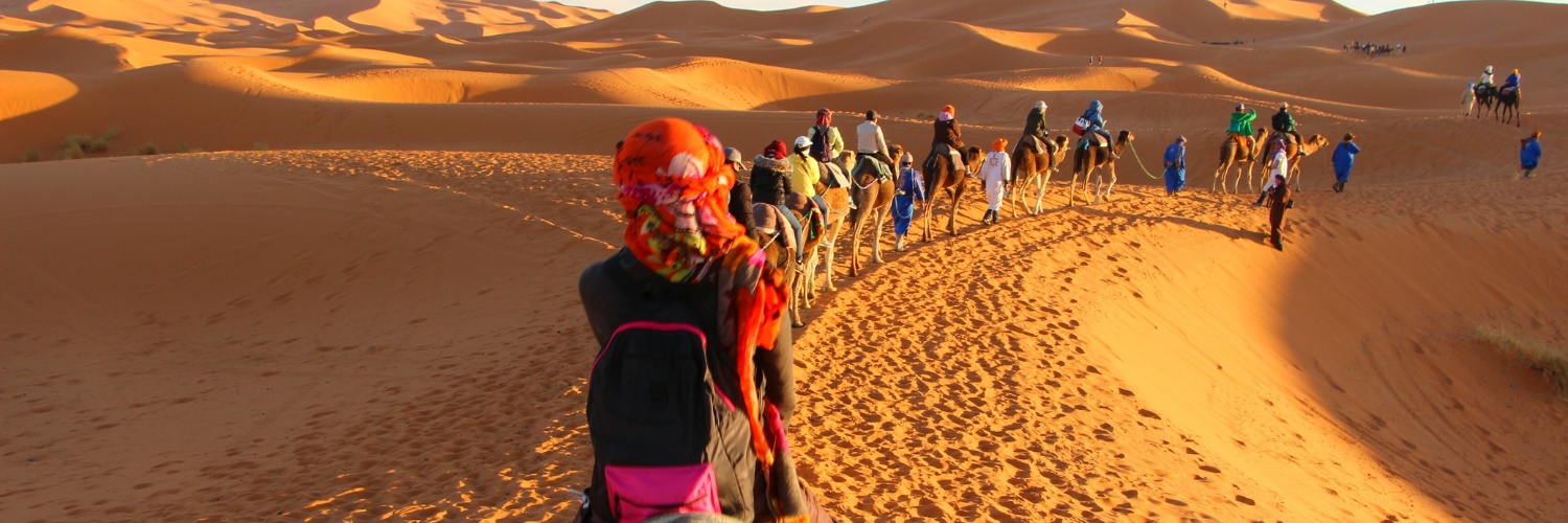 People journeying in the desert, riding atop camels against a backdrop of golden sand dunes.